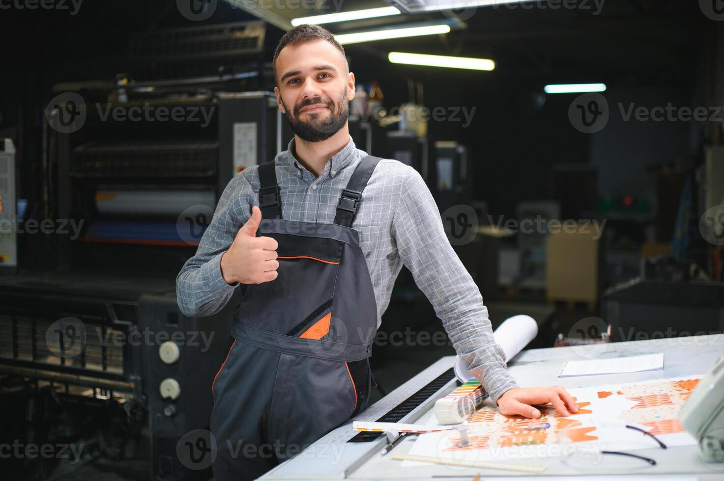 Printing house interior with modern offset print machine and operator in working uniform checking quality and controlling process of print photo