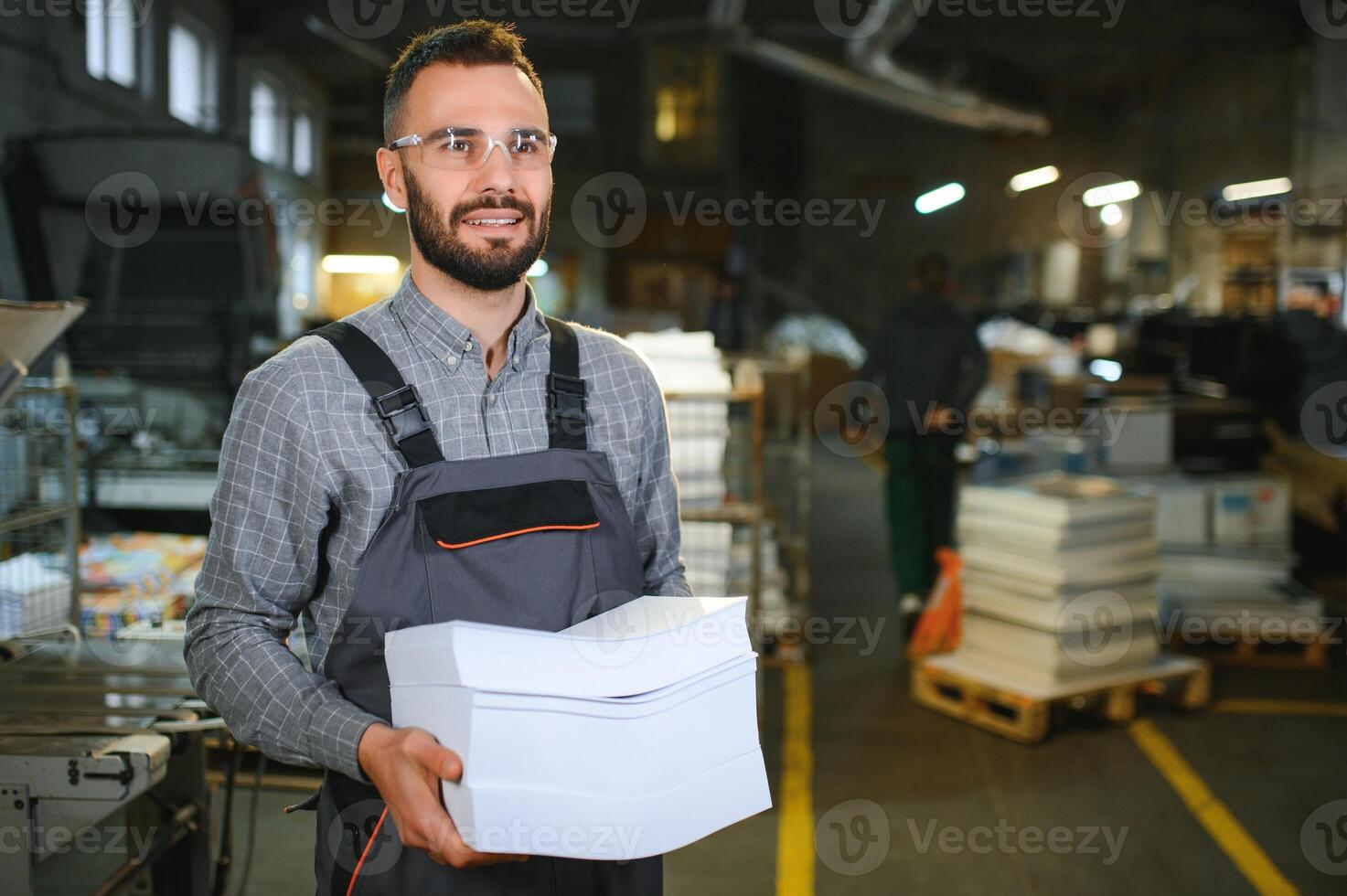 Printing house interior with modern offset print machine and operator in working uniform checking quality and controlling process of print photo