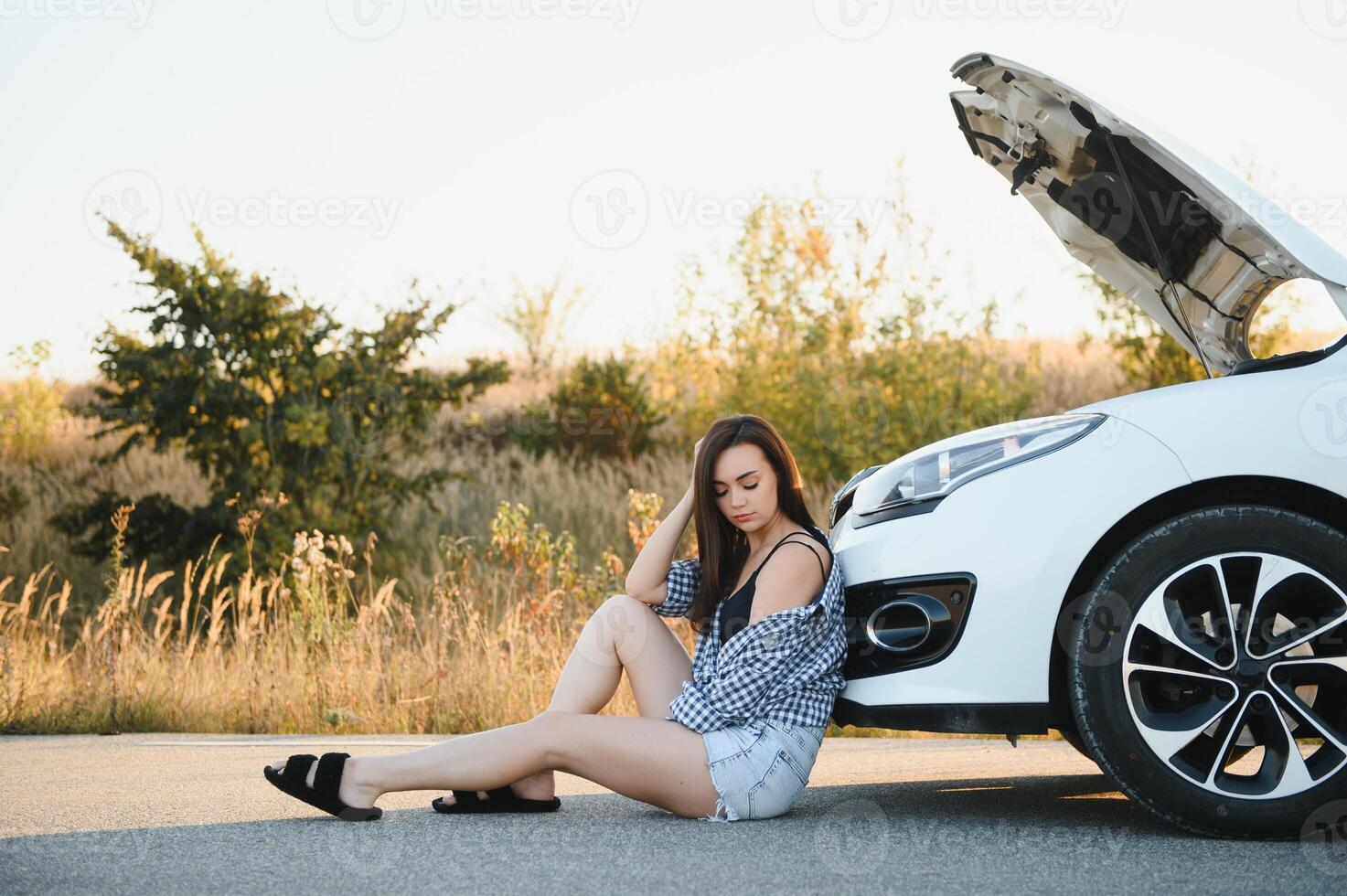 hermosa joven niña sentado a un roto coche en el la carretera desesperado a obtener ayuda foto