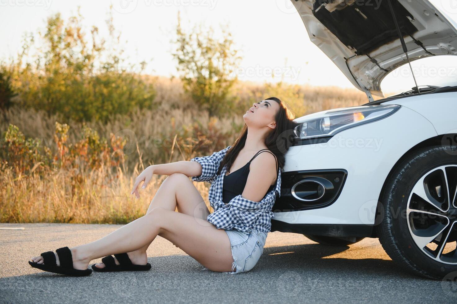 hermosa joven niña sentado a un roto coche en el la carretera desesperado a obtener ayuda foto