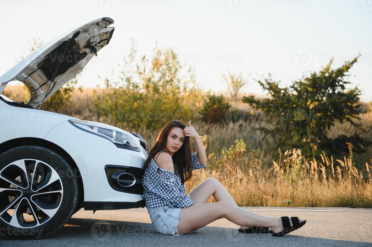 A young girl sits near a broken car on the road with an open hood. photo