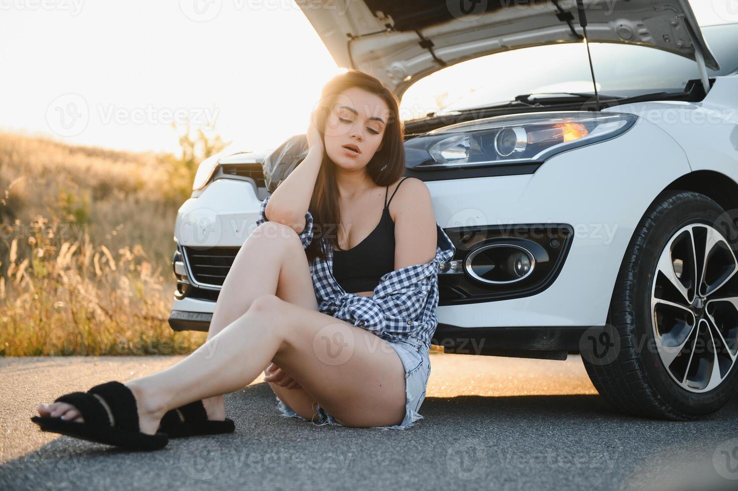 A young girl sits near a broken car on the road with an open hood. photo