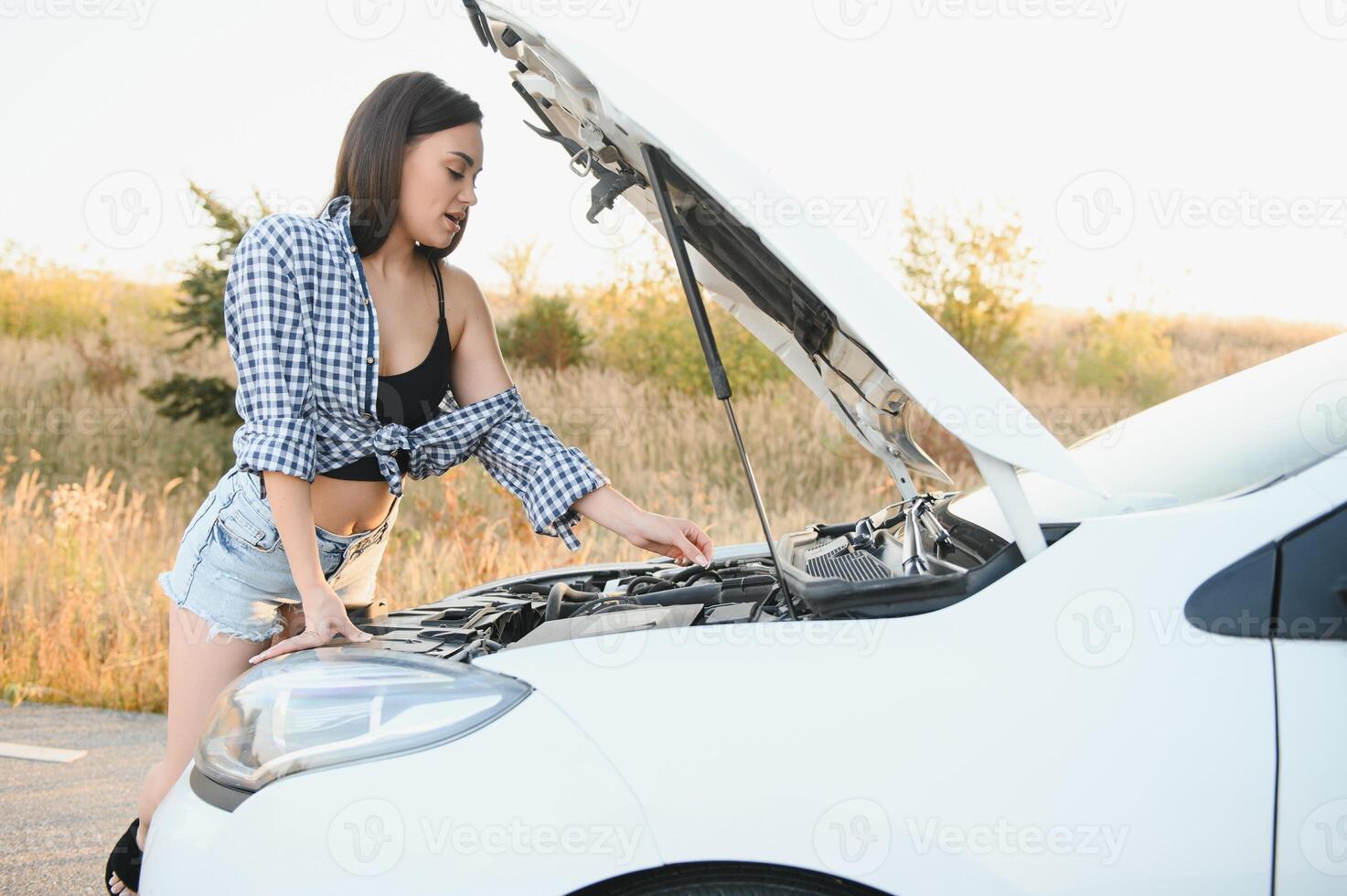 hermosa Delgado niña en camisa y pantalones cortos mira en abierto coche capucha en un la carretera foto