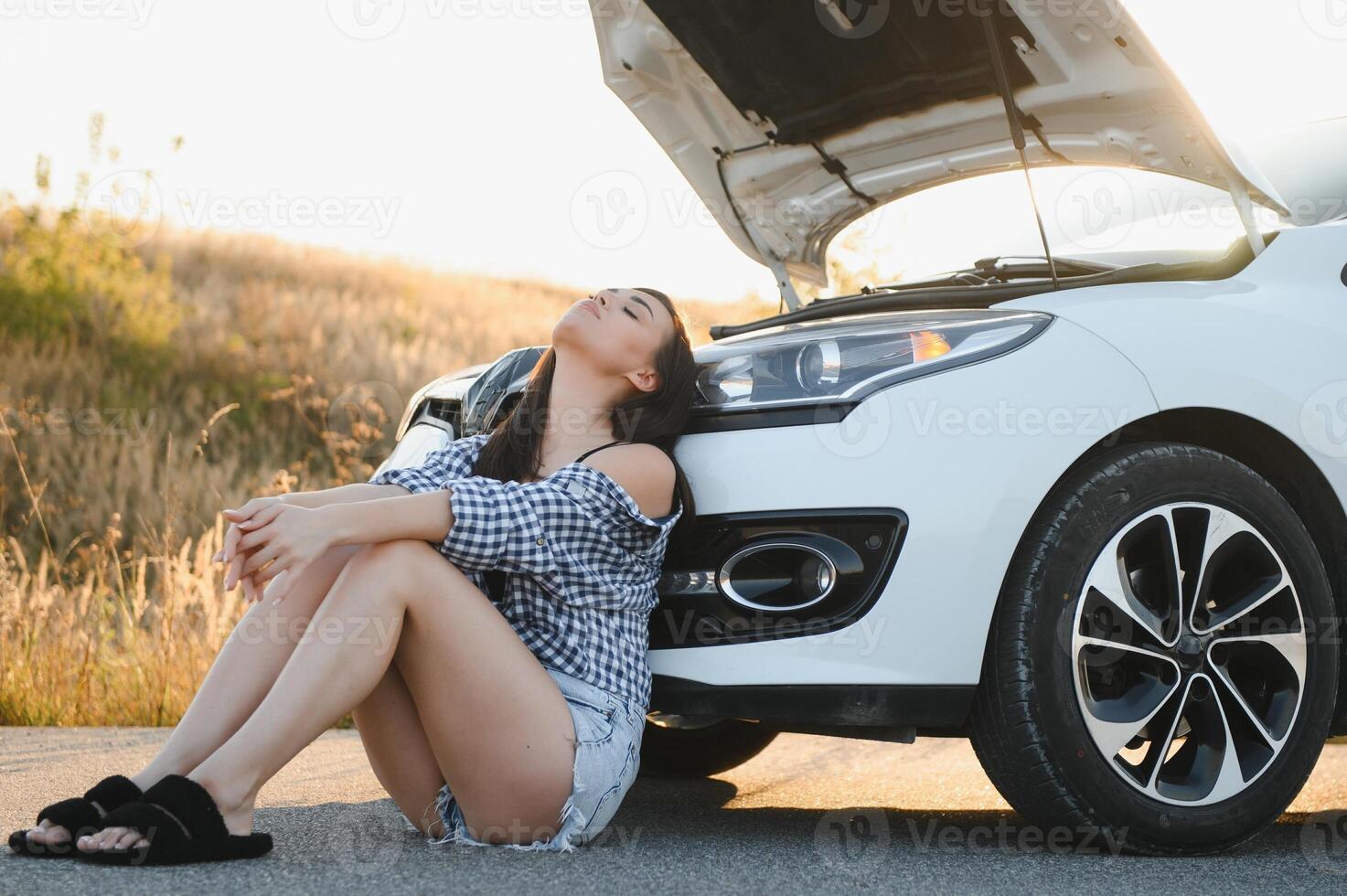 hermosa joven niña sentado a un roto coche en el la carretera desesperado a obtener ayuda foto