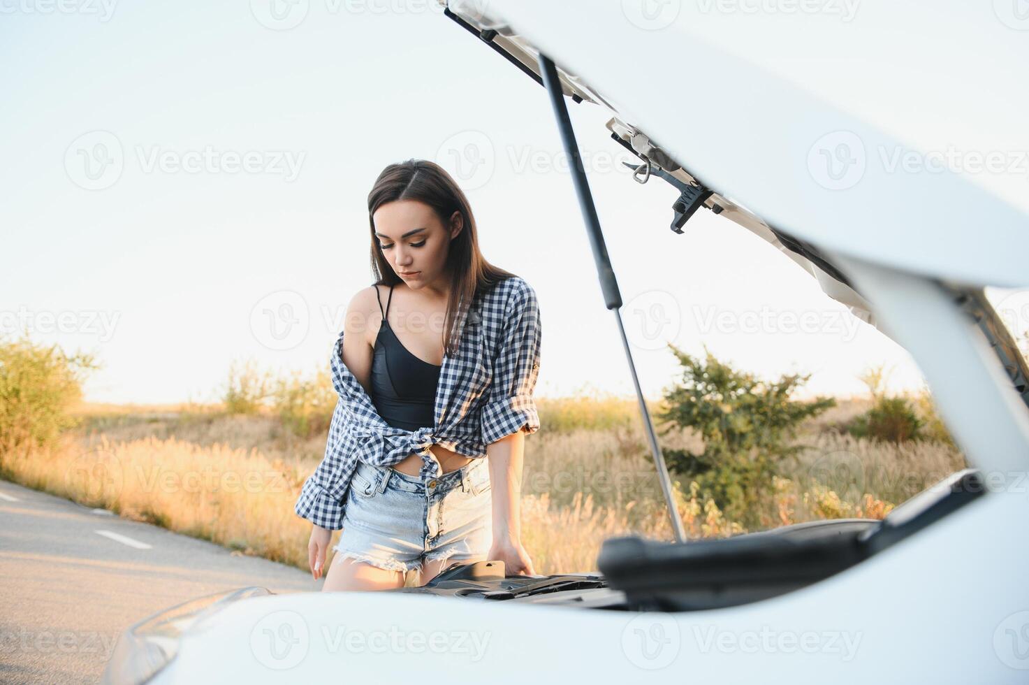 Beautiful slim girl in shirt and shorts looks in open car hood on a road photo