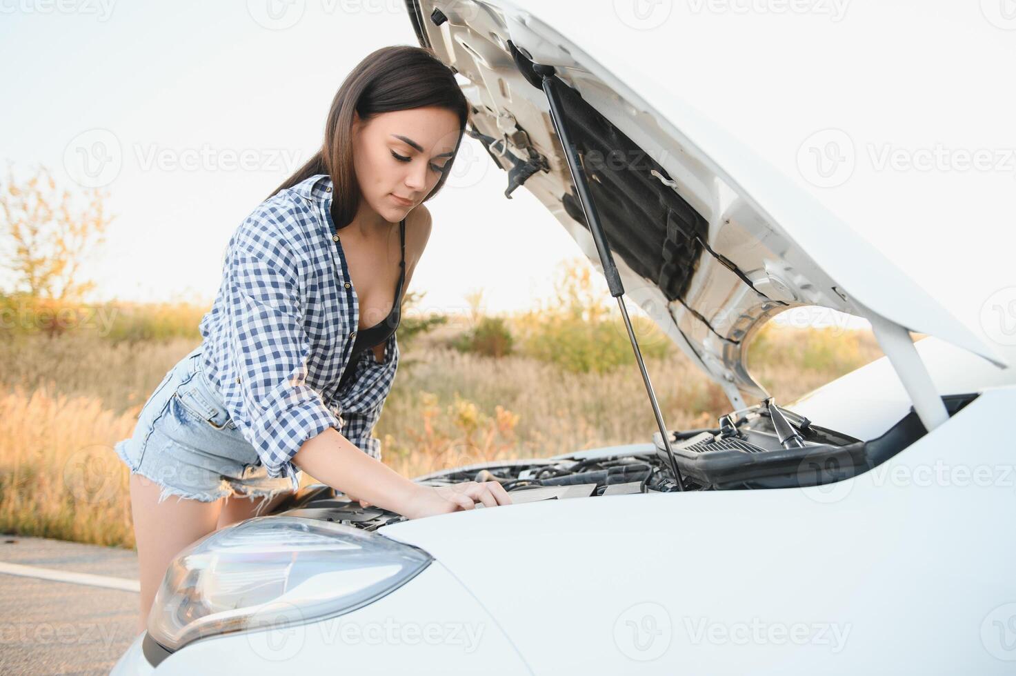 atractivo Delgado joven niña en verano pantalones cortos y camisa refacción un roto coche. un hermosa mujer soportes cerca elevado coche capucha. foto