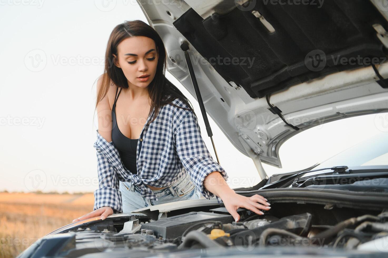 atractivo Delgado joven niña en verano pantalones cortos y camisa refacción un roto coche. un hermosa mujer soportes cerca elevado coche capucha. foto