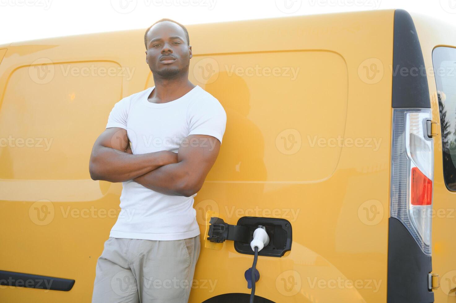 A african american man stands next to yellow electric delivery van at electric vehicle charging stations photo