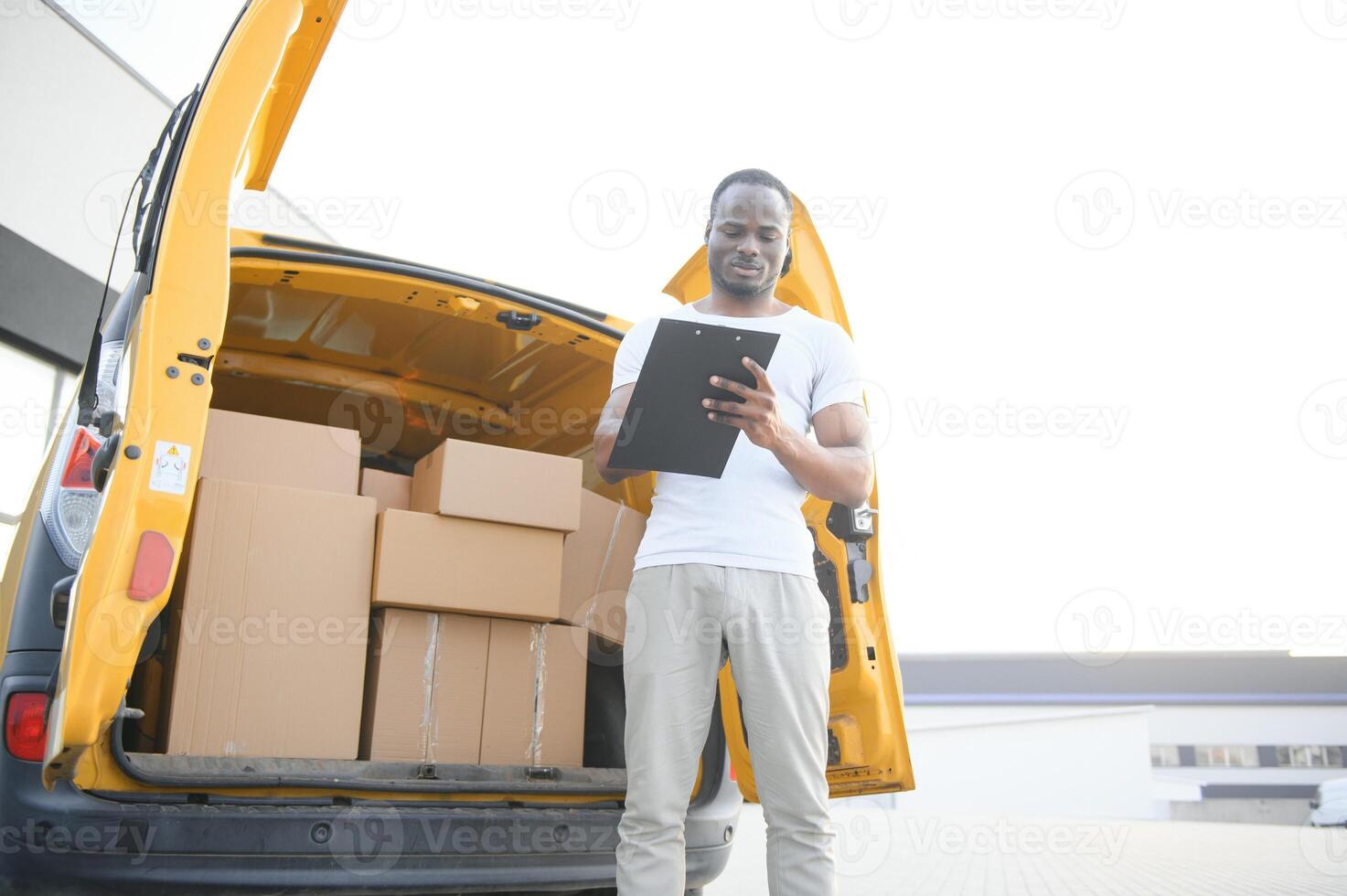african american Worker unloading boxes from van outdoors. Moving service concept. Male employee of moving company makes inventory of packed boxes and writes data to clipboard. photo