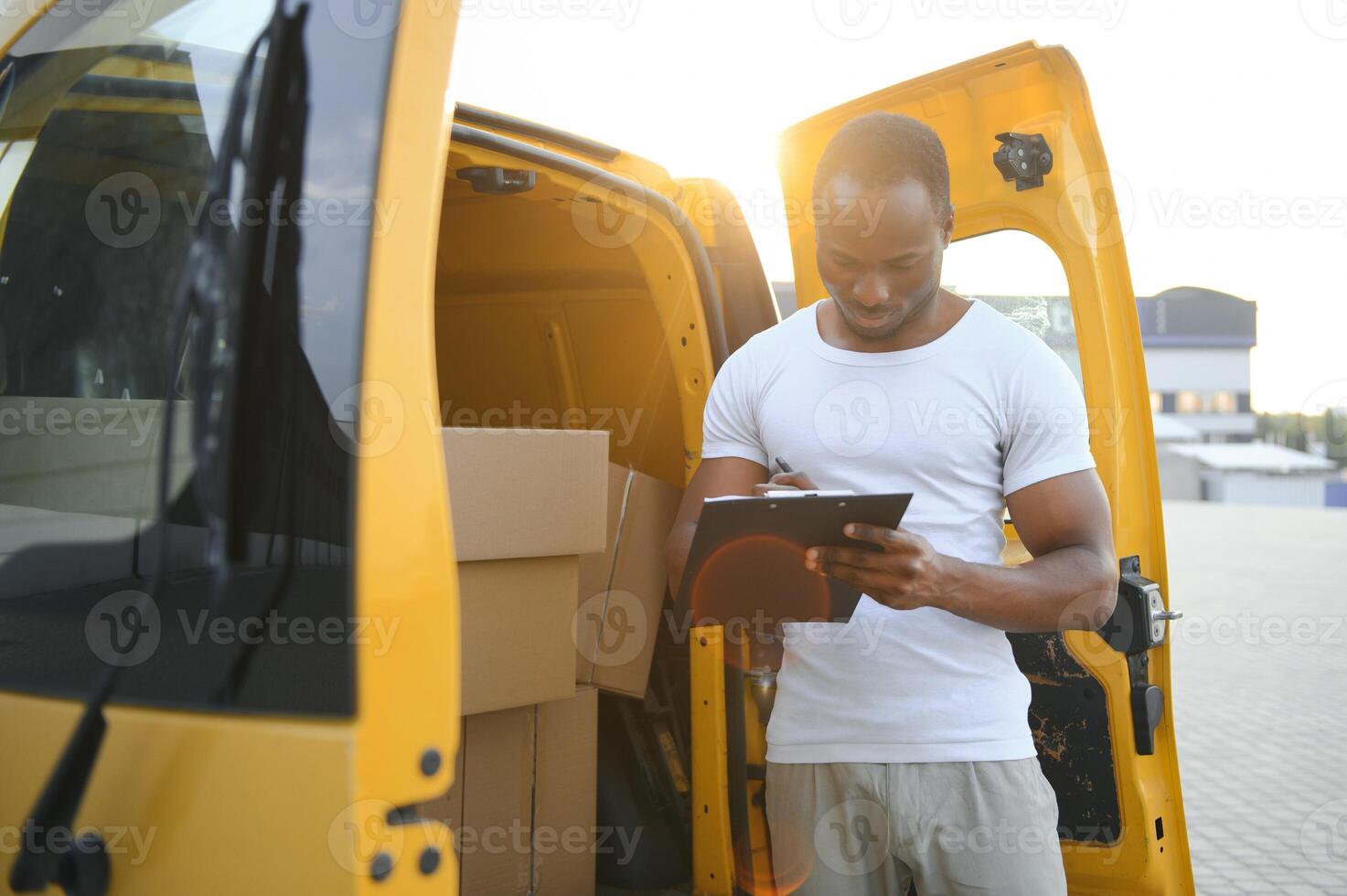 african american Worker unloading boxes from van outdoors. Moving service concept. Male employee of moving company makes inventory of packed boxes and writes data to clipboard. photo