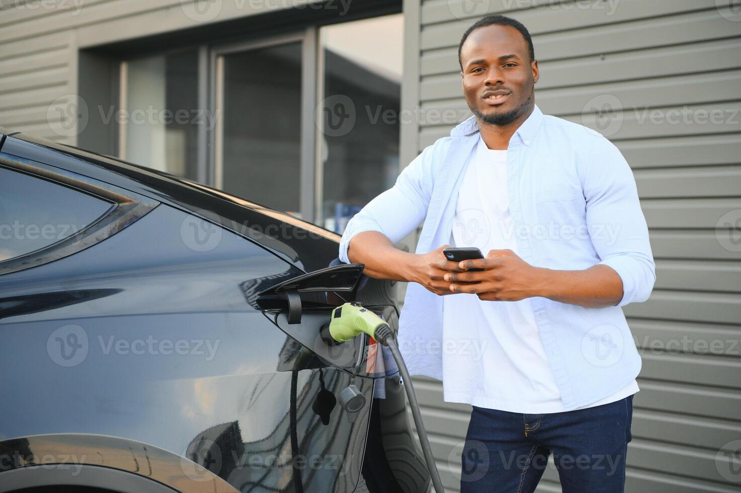 An African man is standing near an electric car, waiting for it to charge at a charging station and using the phone. photo