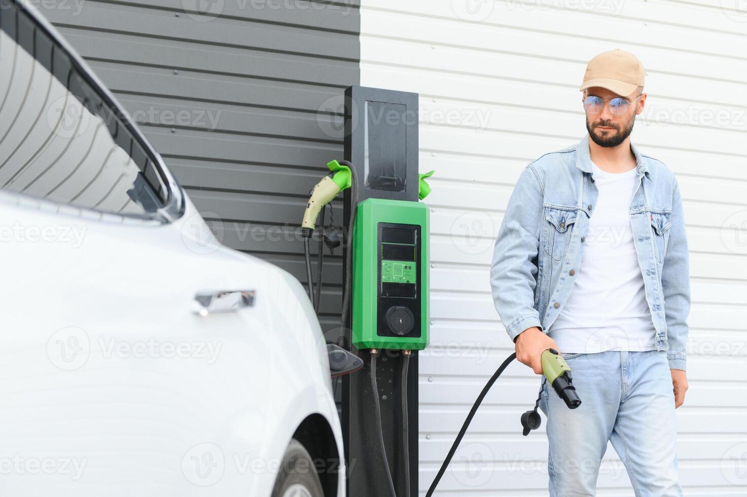 Man Holding Power Charging Cable For Electric Car In Outdoor Car Park. And he s going to connect the car to the charging station in the parking lot near the shopping center. photo