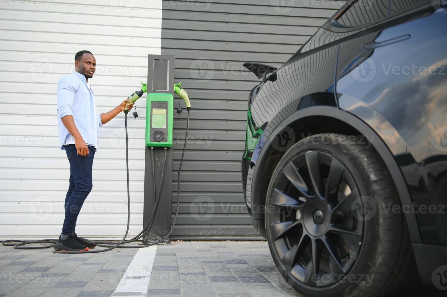 Serious african man holding charge cable in on hand, standing near luxury electric car. photo