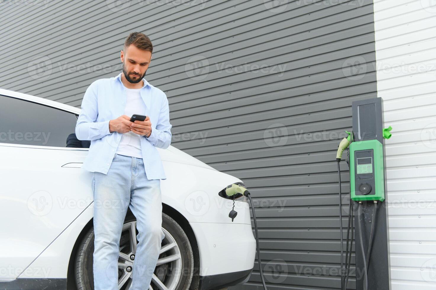 Casual man with smartphone near electric car waiting for the finish of the battery charging process photo