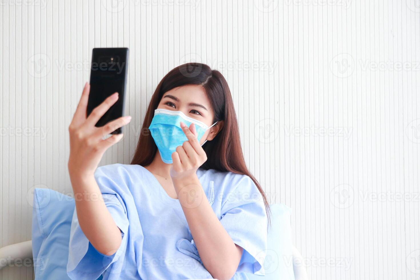 Asian female patient wearing mask in hospital patient room She holds a smartphone online talking to her family. Concept of medical services. patients infected with coronavirus. copy space photo