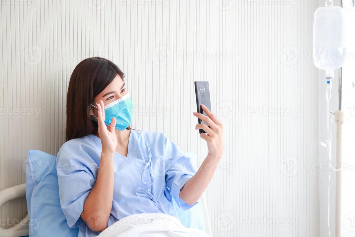 Asian female patient wearing mask in hospital patient room She holds a smartphone online talking to her family. Concept of medical services. patients infected with coronavirus. copy space photo