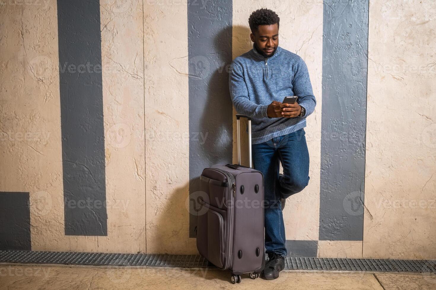 contento hombre con maleta utilizando teléfono mientras propensión en contra un pared en paso de el ferrocarril estación. Copiar espacio en imagen para tu Propaganda o texto. foto