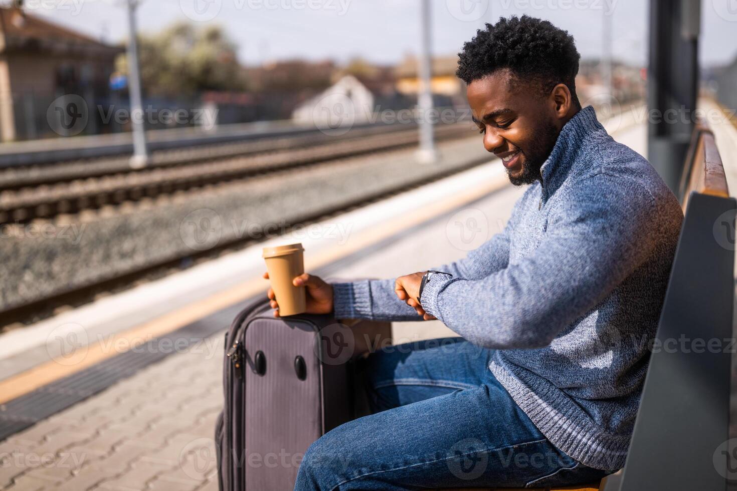 contento hombre con maleta y café mirando un reloj mientras sentado en un banco a el ferrocarril estación. foto