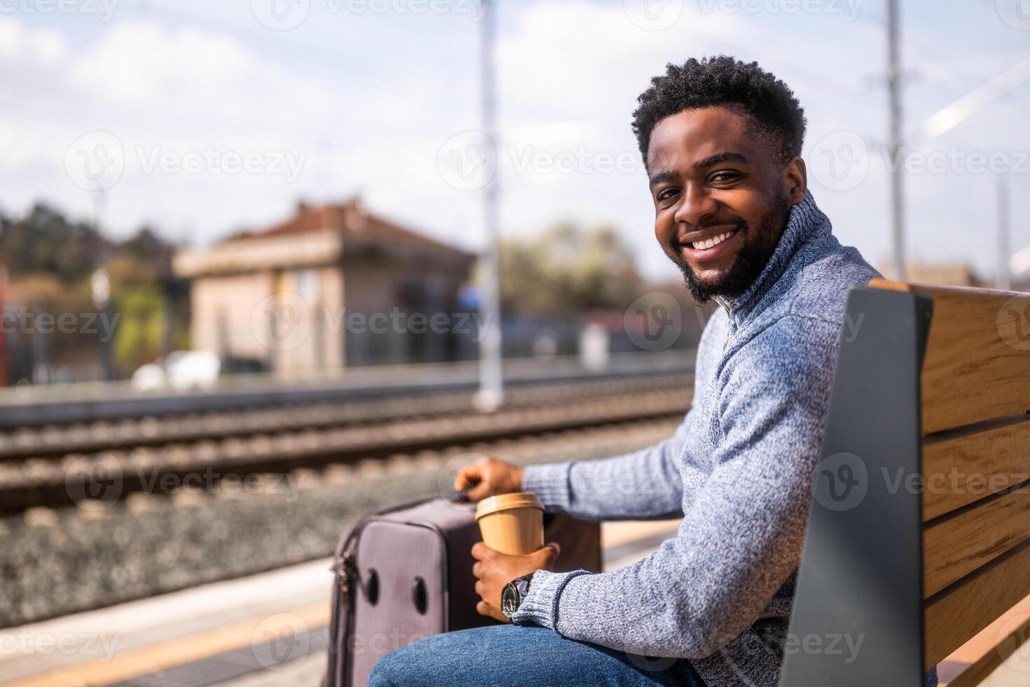 Happy man with suitcase enjoys drinking coffee while sitting on a bench at the railway station. photo