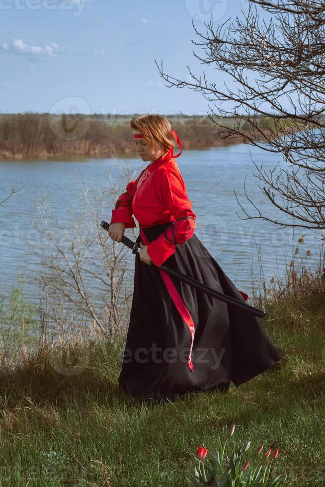 un mujer con corto rojo pelo en negro y rojo hanfu y un katana en el antecedentes de un río en primavera foto