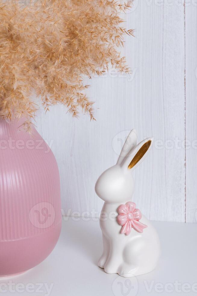 A white porcelain rabbit with a pink bow on a shelf next to a pink vase and a reed photo