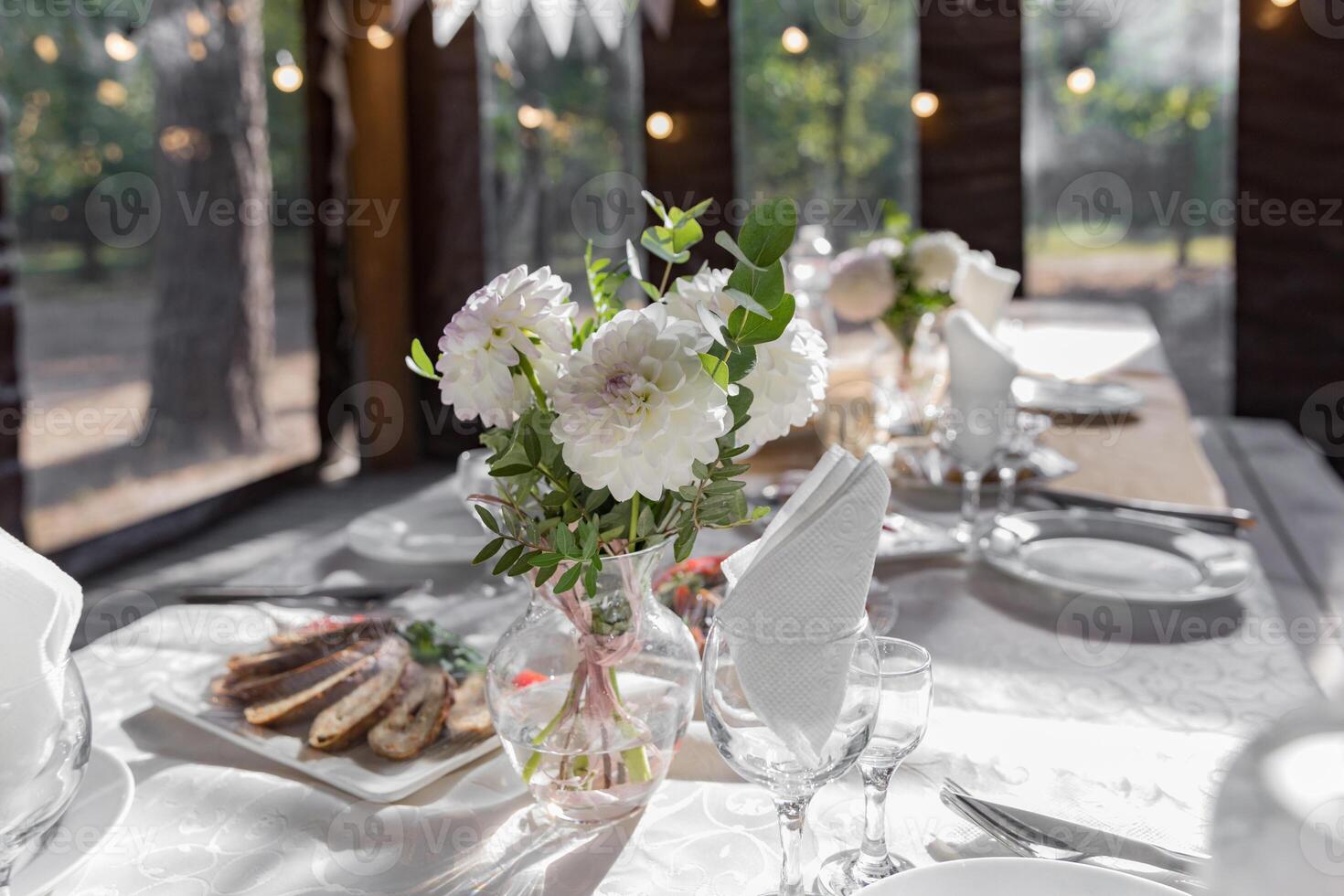 A long festive table decorated with white flowers and greenery. There are plates, glasses and candles on the table. The room is decorated with white garlands. photo