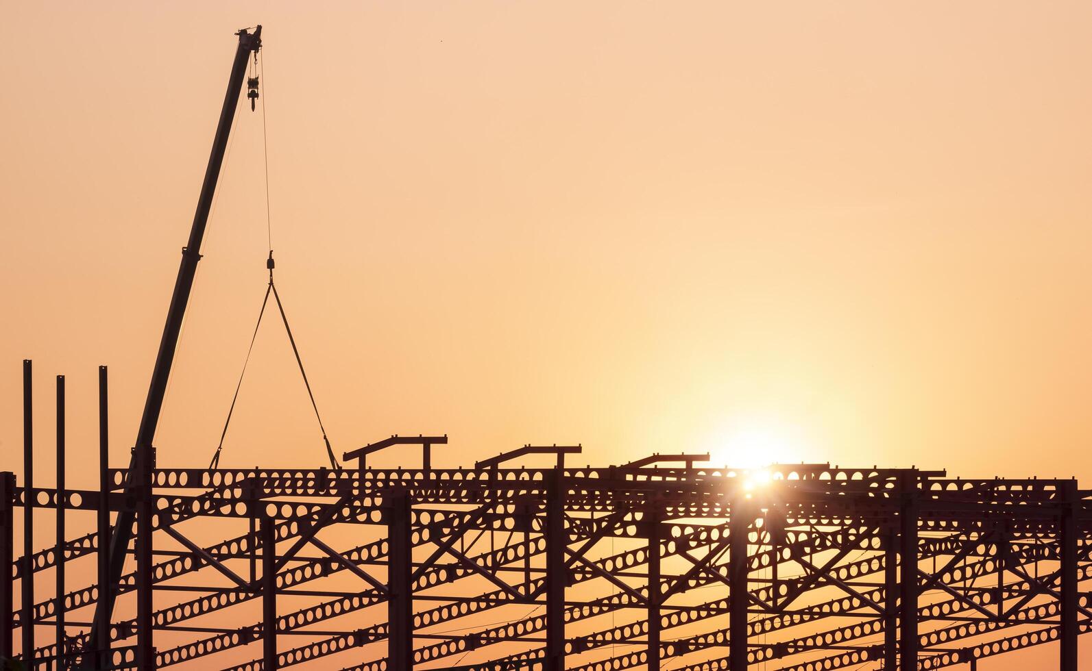 Silhouette crane lifting metal castellated beam to installing on large industrial building structure in construction site area against sunset sky background photo