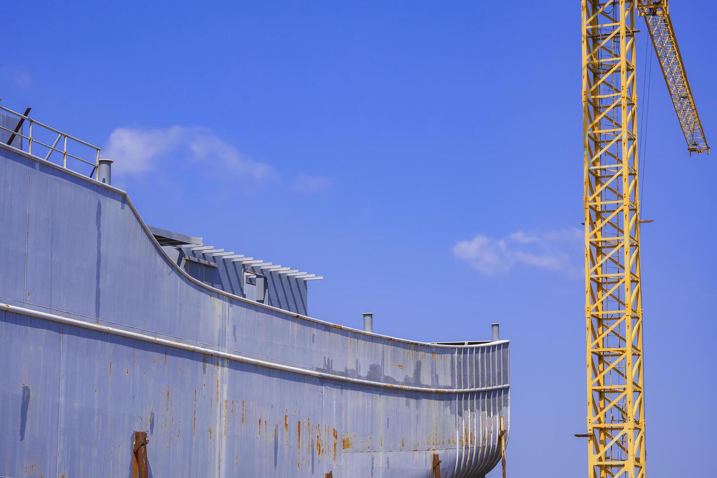 Side view of metal vessel hull during maintenance work in dry dock with tower crane against blue sky in shipyard area photo