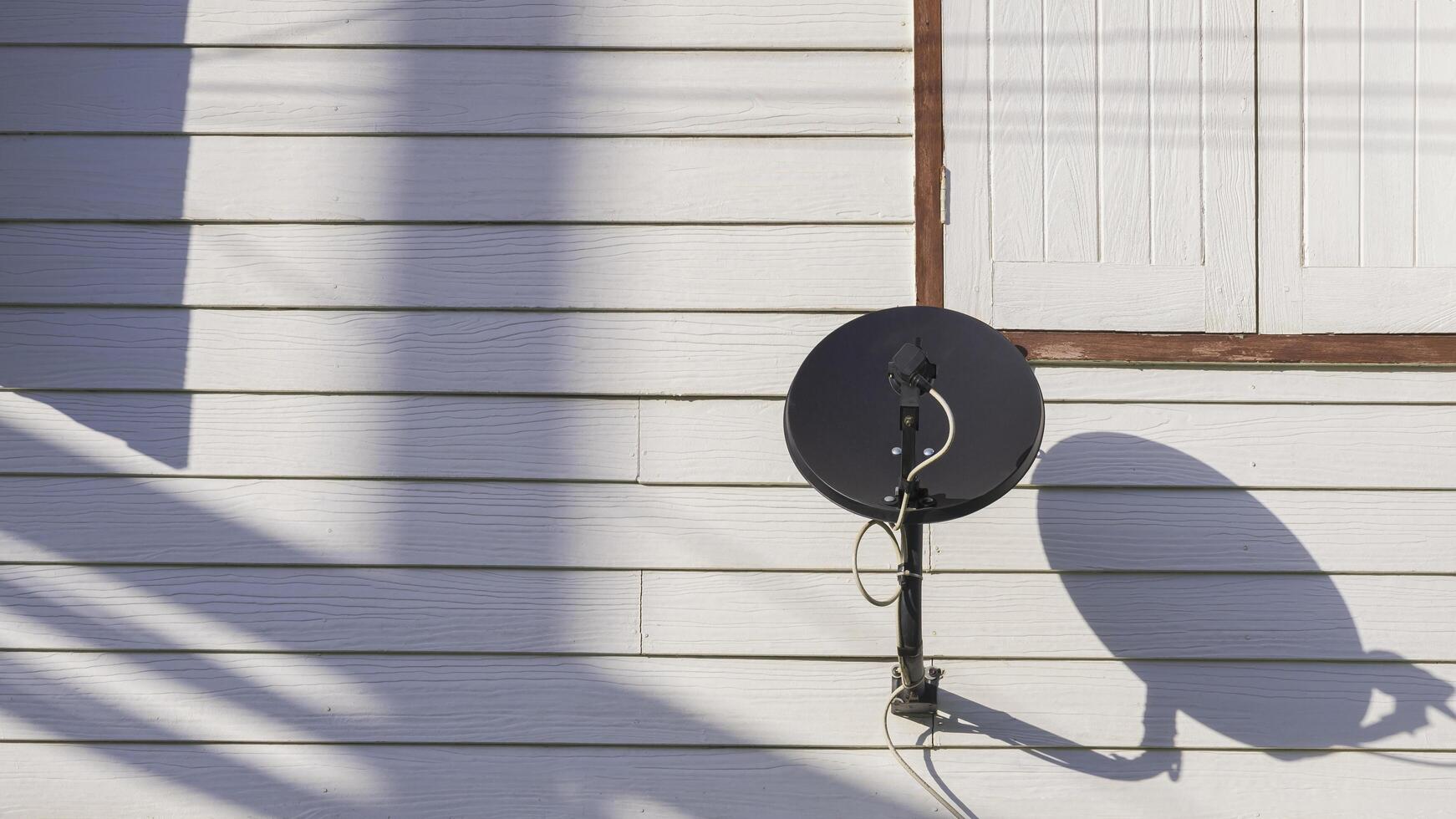 Black satellite dish on white artificial wooden wall of vintage house with sunlight and shadow on surface photo