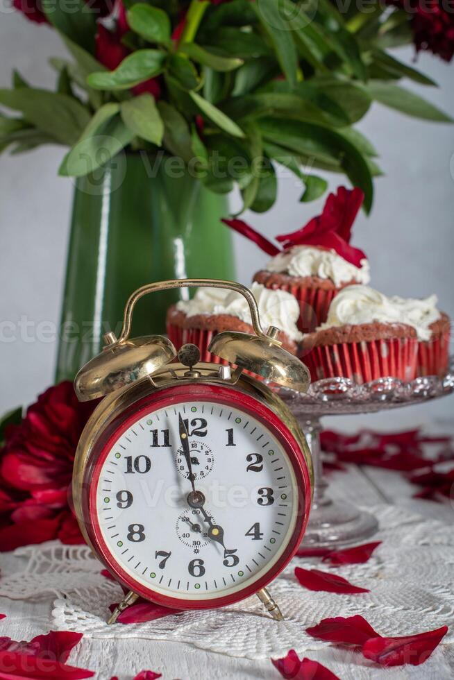 todavía vida en Inglés estilo con escarlata peonías y rojo terciopelo magdalenas con en un plato, té hora en rojo antiguo reloj foto