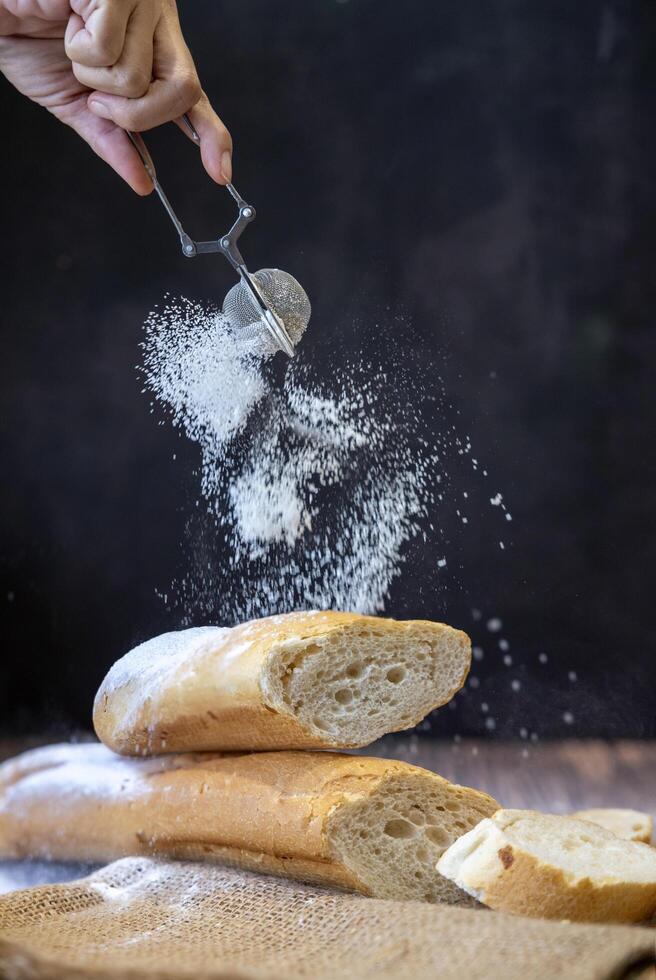 Hand of baker dusting the baguette bread with icing sugar on the dark black background for pastry and bakery concept photo