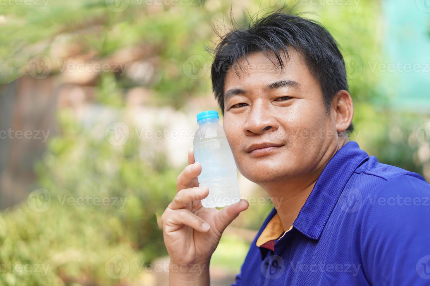 Handsome Asian man hold cool drinking water bottle to touch his cheek for relief hot weather condition. Concept, self first aid to protect from heat stroke, cool down. Health care. photo