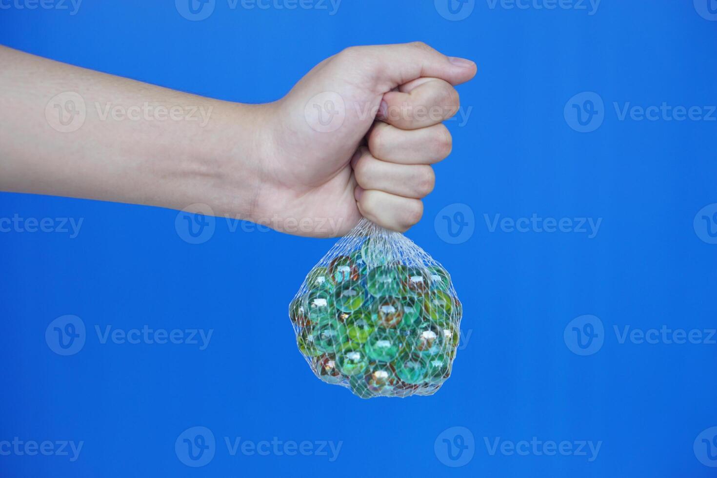 Close up hand hold net bag of marbles balls, small spherical object often made from glass. Blue background. Concept. object use as toy for playing games or use for DIY craft decoration. photo