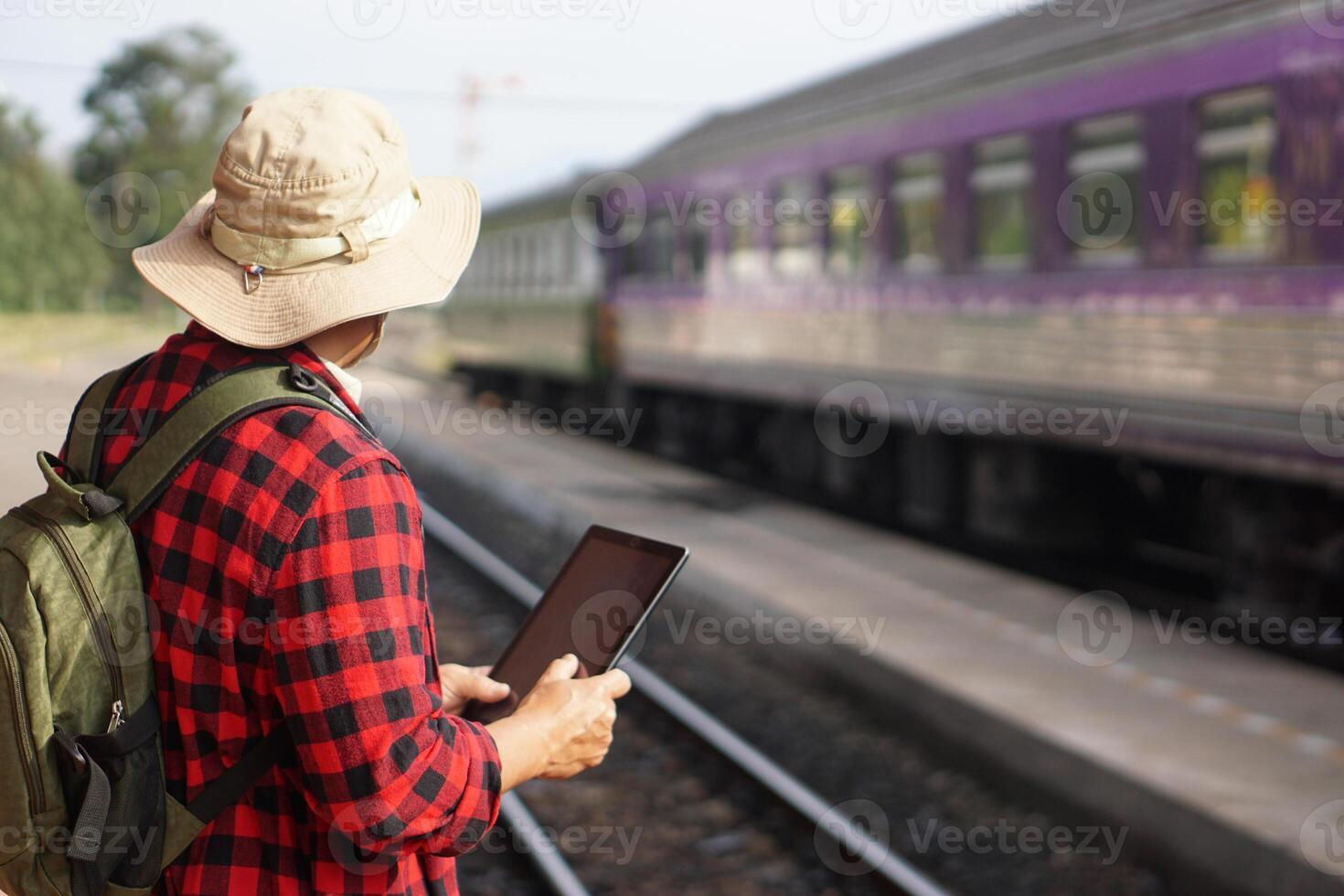 Asian man traveller is at railway station, wears hat, holds smart tablet. Concept, travel by train in Thailand can book or by ticket online. Technology and transportation. photo