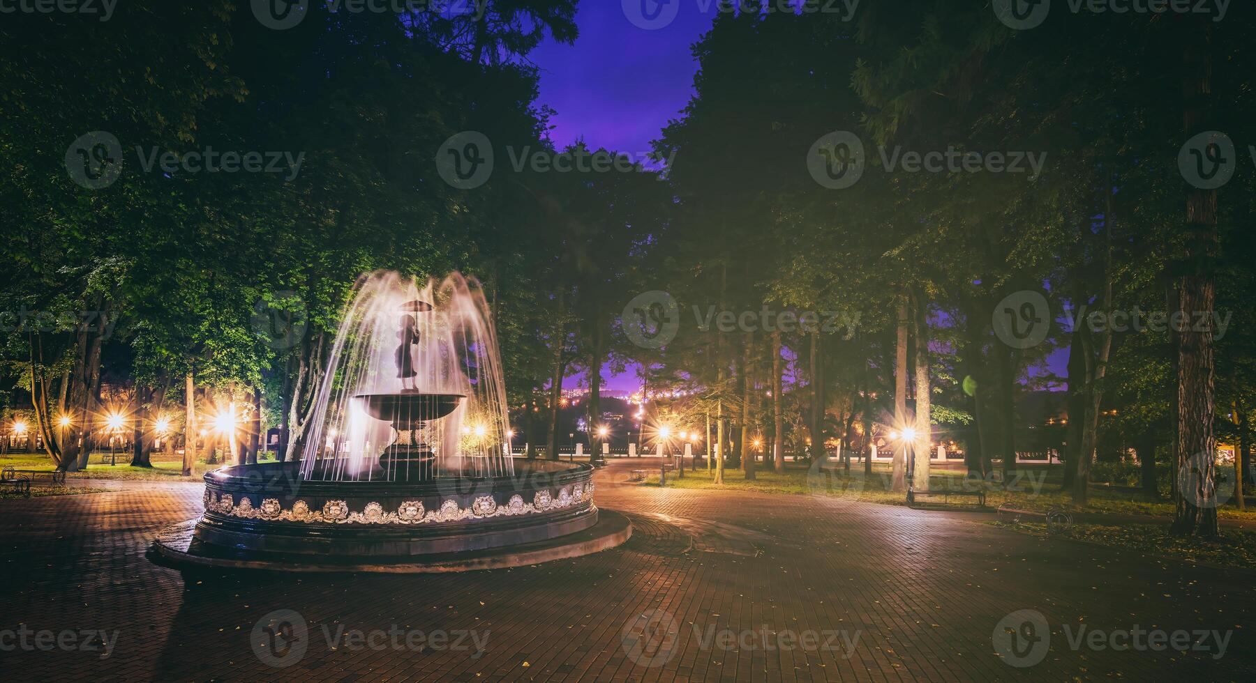 A fountain with blurred streams of water in a night park illuminated by lanterns with a stone pavement, trees and benches. Vintage film aesthetic. photo