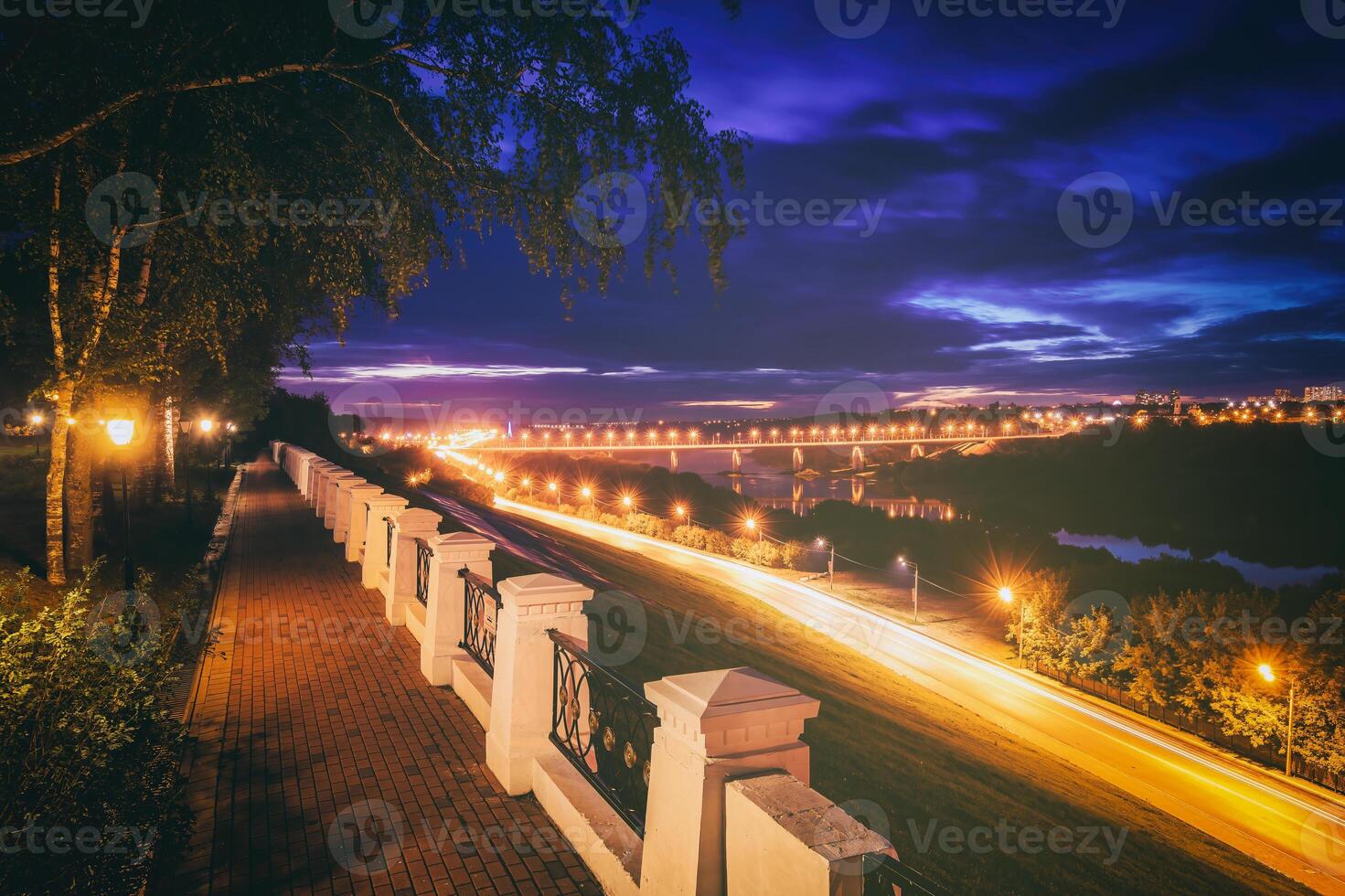Moving car with blur light through city at night. Bridge over the river and the road. A view from the park from a height with a fence in the foreground. Vintage film aesthetic. photo