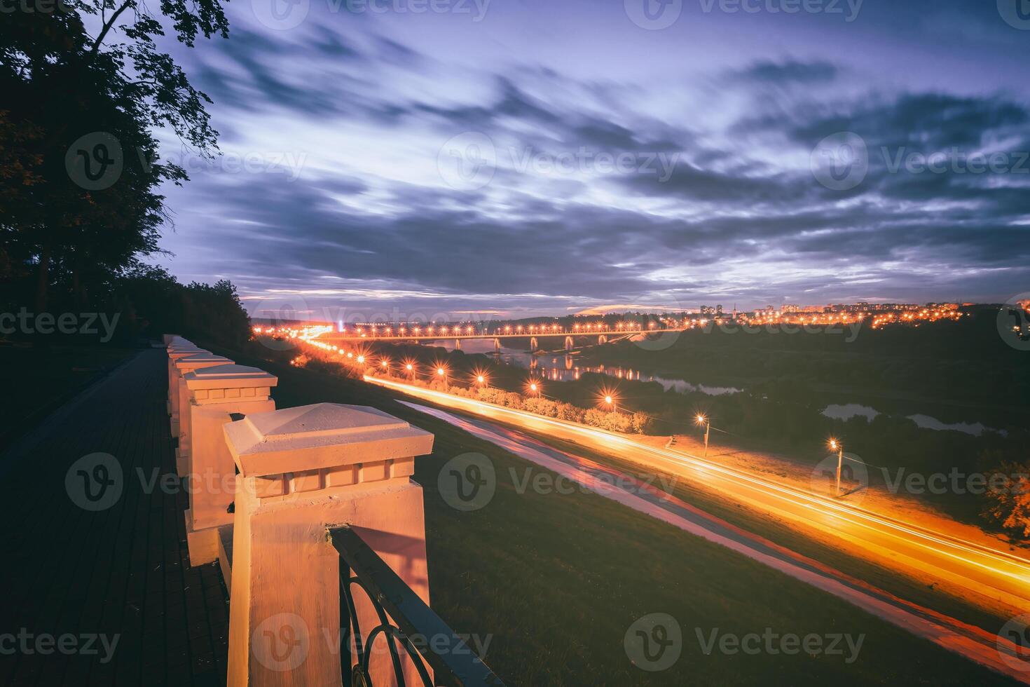 Moving car with blur light through city at night. Bridge over the river and the road. A view from the park from a height with a fence in the foreground. Vintage film aesthetic. photo