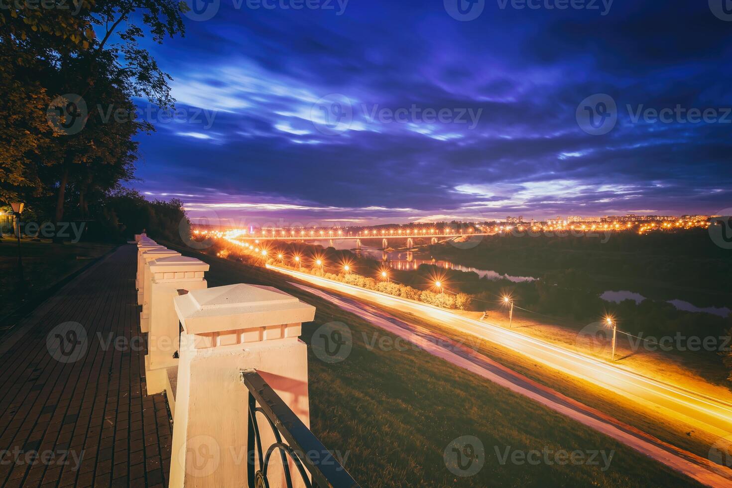 Moving car with blur light through city at night. Bridge over the river and the road. A view from the park from a height with a fence in the foreground. Vintage film aesthetic. photo