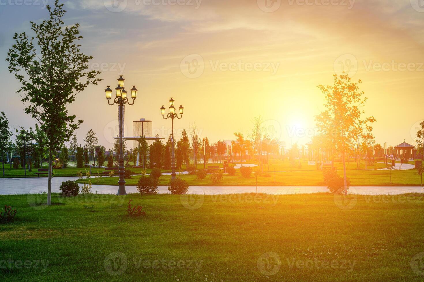 City park in early summer or spring with lanterns, young green lawn, trees and dramatic cloudy sky on a sunset or sunrise. photo