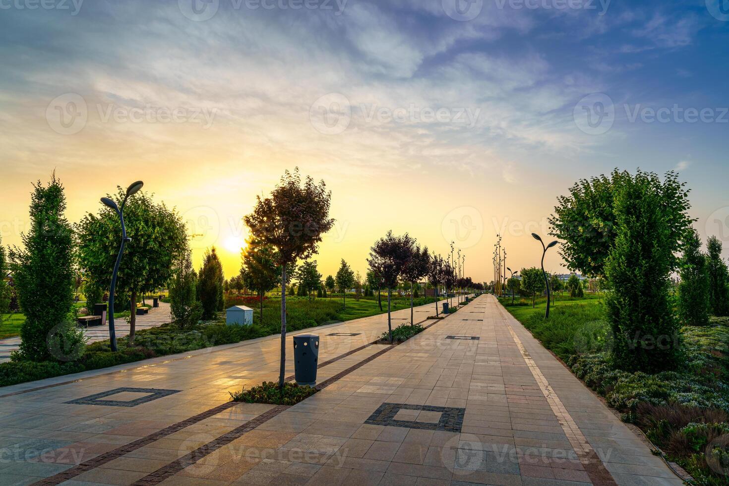 City park in early summer or spring with pavement, lanterns, young green lawn, trees and dramatic cloudy sky on a sunset or sunrise. photo