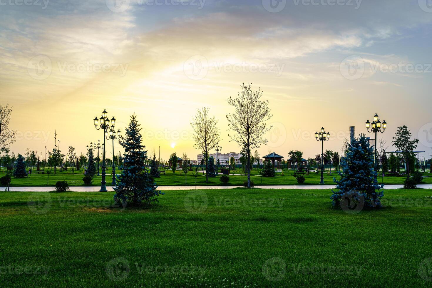 City park in early summer or spring with lanterns, young green lawn, trees and dramatic cloudy sky on a sunset or sunrise. photo