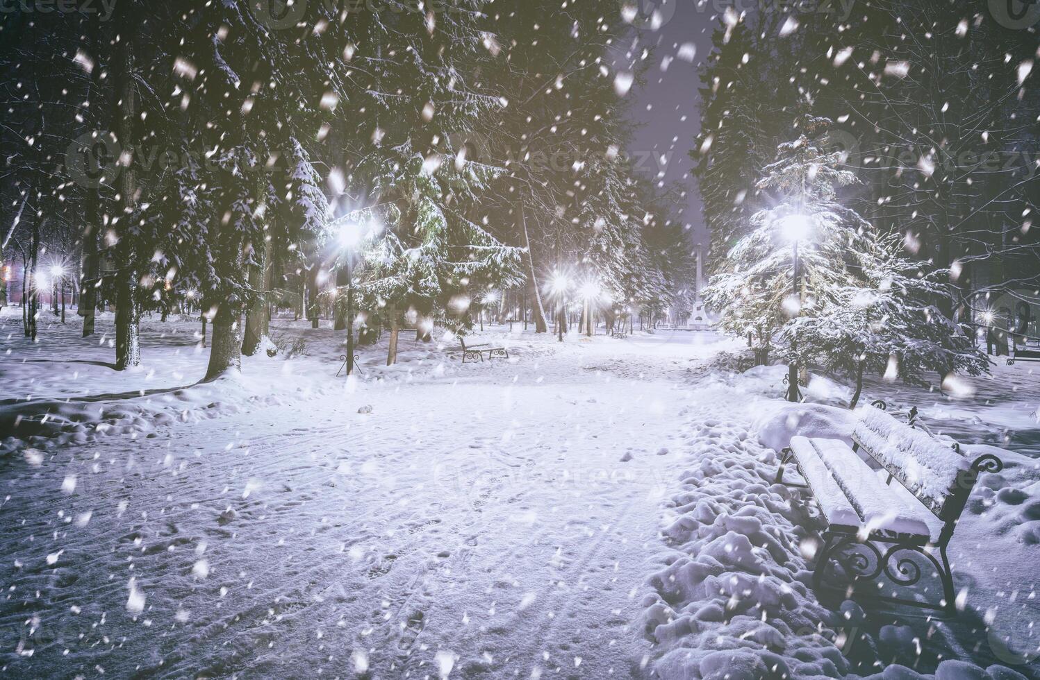 Snowfall in a winter park at night with glowing lanterns, pavement covered with snow and trees. Vintage film aesthetic. photo