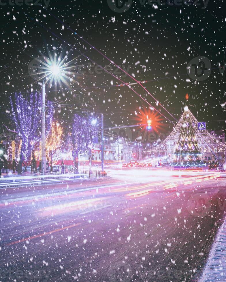 Night winter city with Christmas or New Year decorations, spruce and traces of headlights of moving cars in a snowfall. Vintage film aesthetic. photo