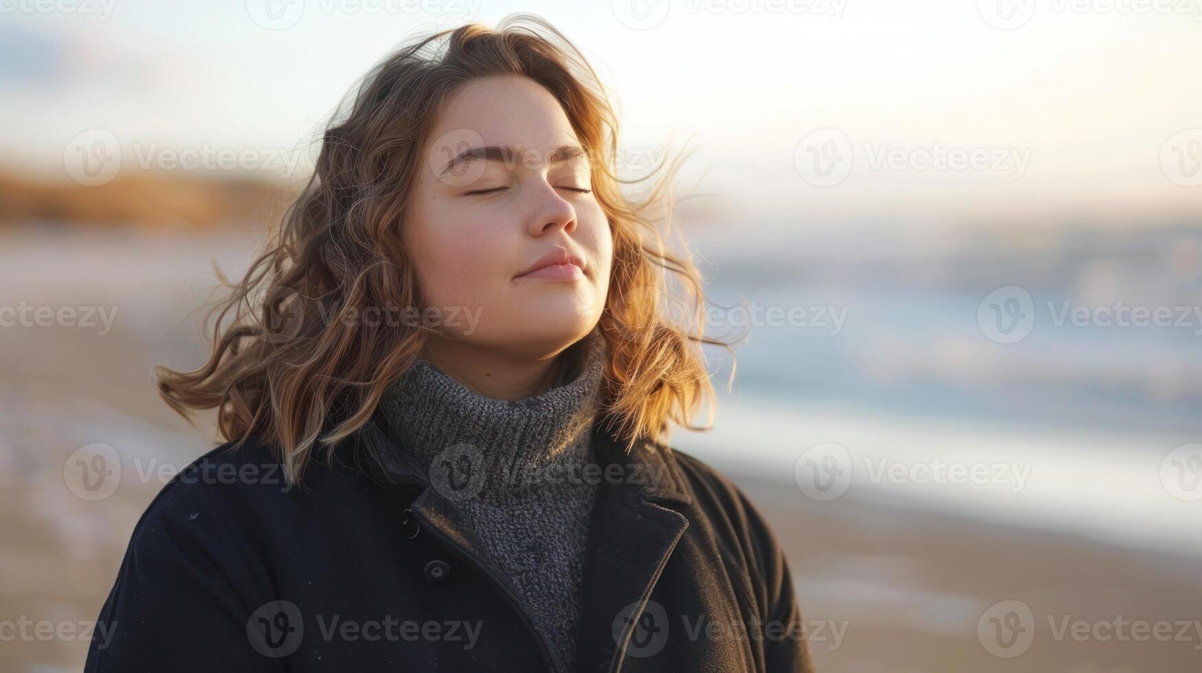 A woman in casual attire standing on a sandy beach, eyes closed under the sunlight photo