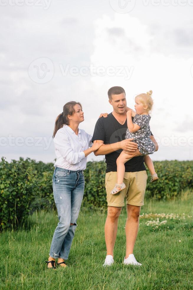 The daughter hugging parents on nature. Mom, dad and girl toddler, walk in the grass. Happy young family spending time together, outside, on vacation, outdoors. The concept of family holiday photo