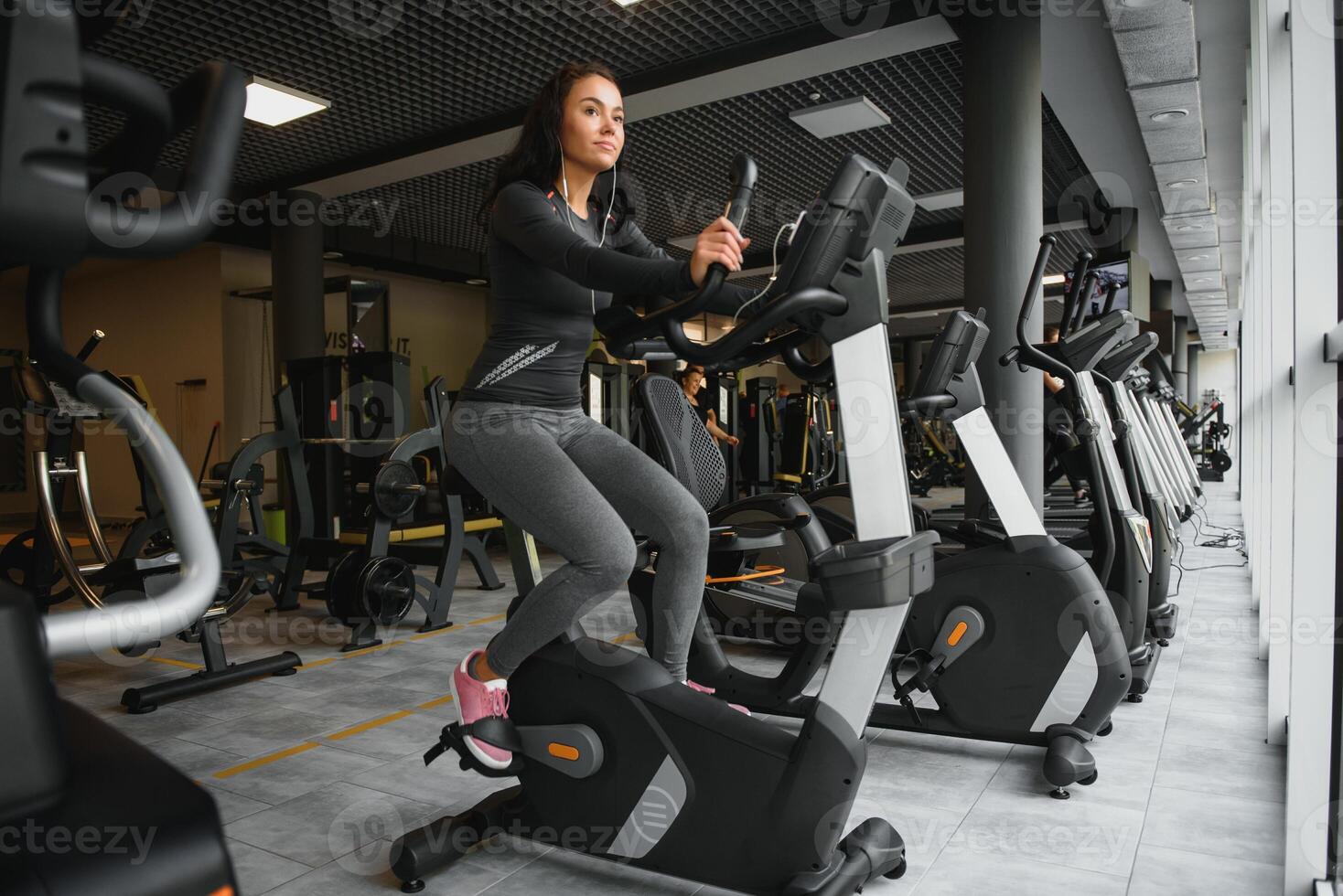 joven mujer con auriculares haciendo ejercicios en papelería bicicleta en un gimnasio o aptitud centro. joven deportivo mujer en gimnasio escucha música desde teléfono inteligente mujer haciendo cardio ejercicios foto