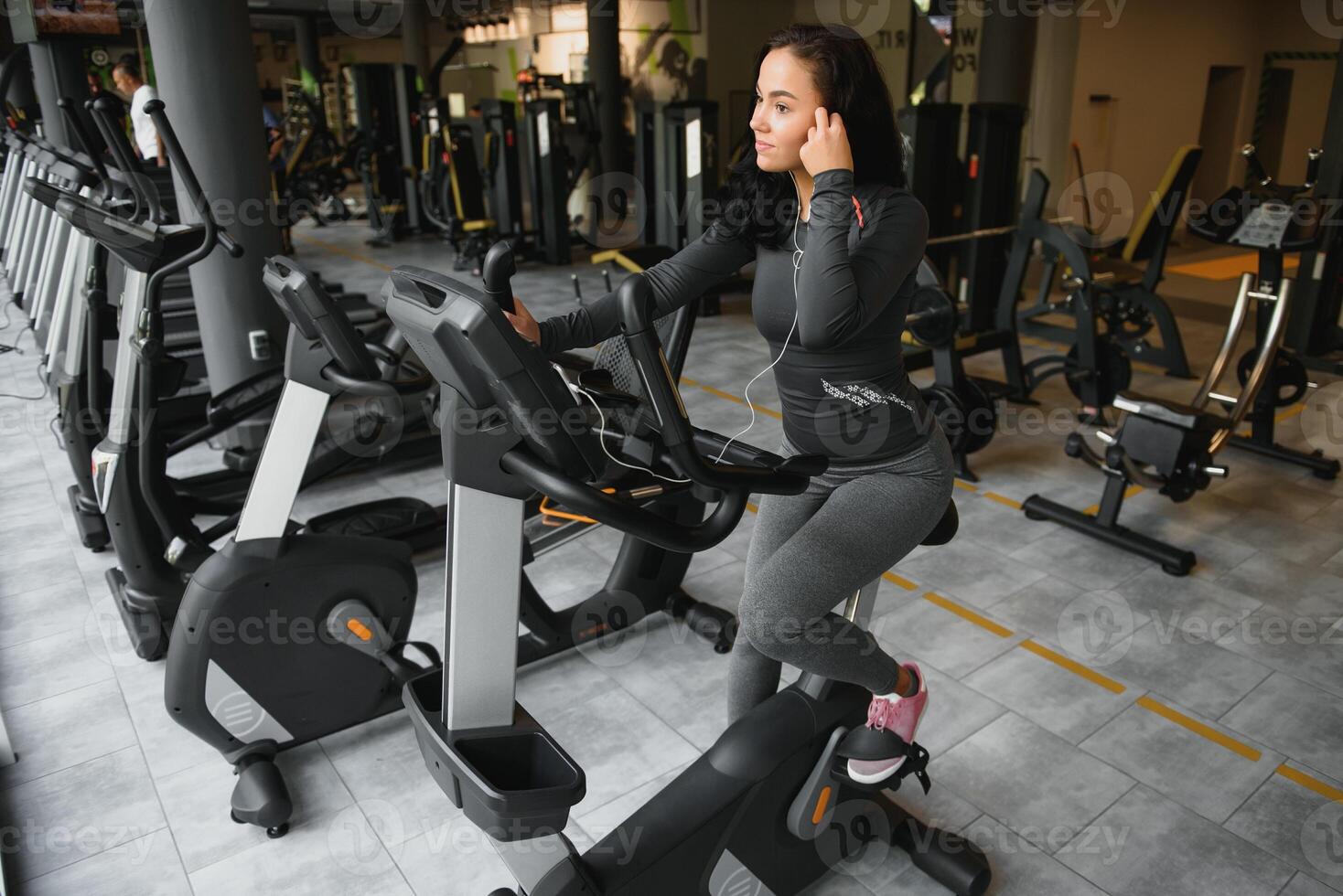 Young woman with headphones doing exercises on stationery bicycle in a gym or fitness center. Young sporty woman in gym listen music from smartphone. Women doing cardio exercises photo