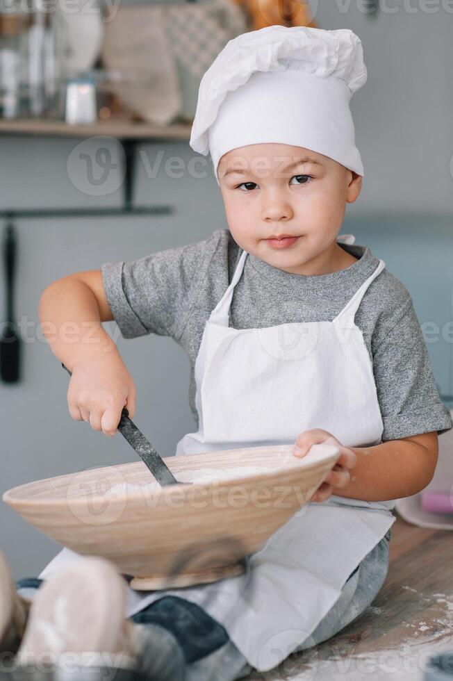 joven chico linda en el cocina cocinar cocinero en blanco uniforme y sombrero cerca mesa. hecho en casa pan de jengibre. el chico cocido el chocolate galletas foto