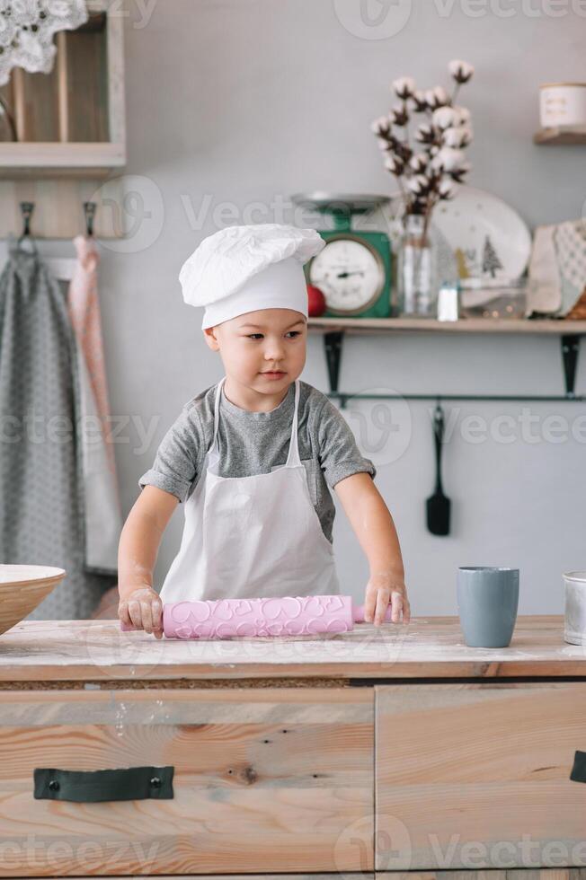 joven chico linda en el cocina cocinar cocinero en blanco uniforme y sombrero cerca mesa. hecho en casa pan de jengibre. el chico cocido el chocolate galletas foto