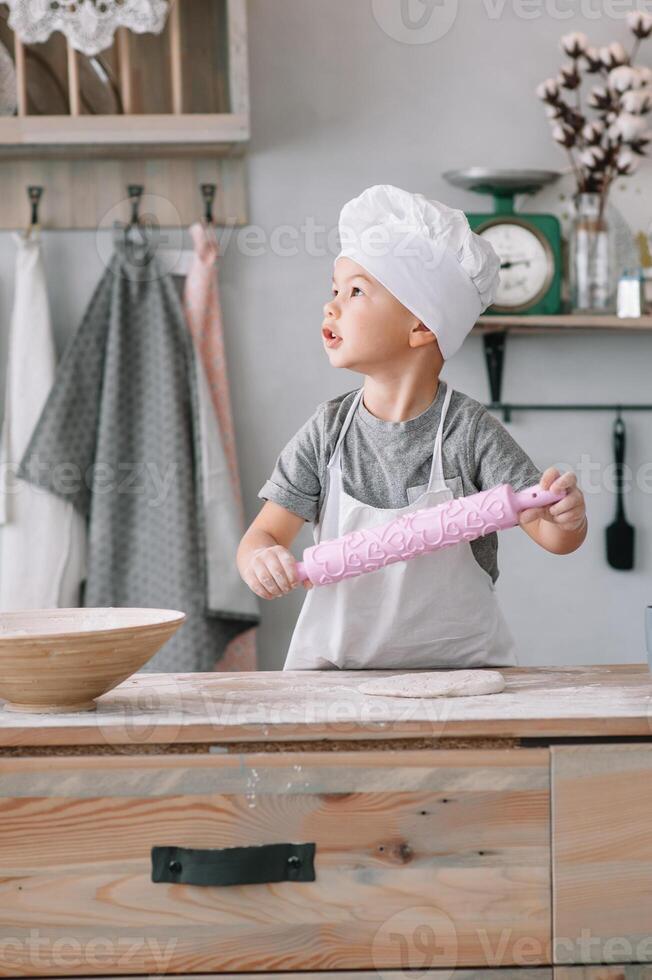 joven chico linda en el cocina cocinar cocinero en blanco uniforme y sombrero cerca mesa. hecho en casa pan de jengibre. el chico cocido el chocolate galletas foto